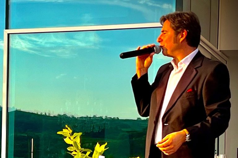 Entertainer im Smoking mit Mikrofon singt im Freien vor einer Glaswand und einer Landschaft im Hintergrund.